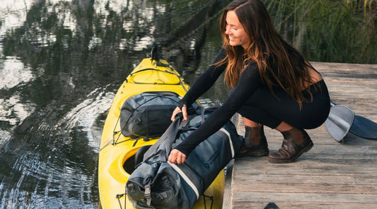 A HAPPY LADY SMILING AS SHE PLACES HER NORTH STORM 60 LITRE DUFFLE BAG INTO A KAYAK.