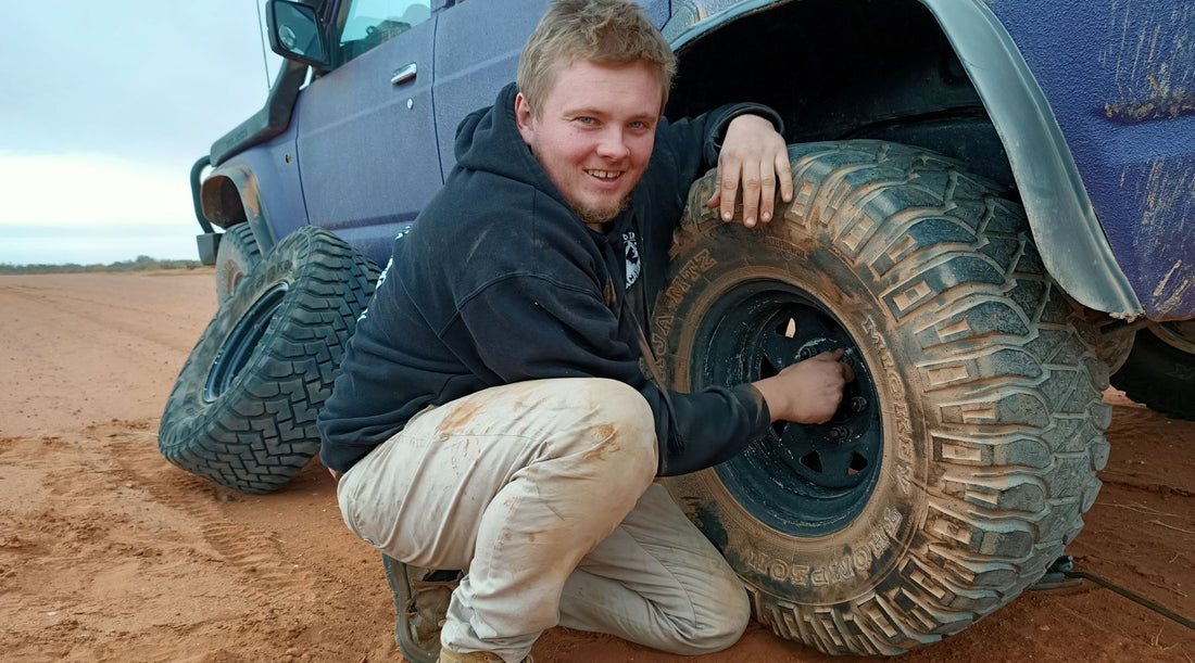 TOMMO CROUCHED BY A 4 WHEEL DRIVE WHEEL SMILING AT THE CAMERA.