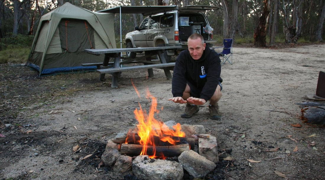 KEV SMITH OF WOOLGOOLGA OFFROAD CROUCHED BY A CAMPFIRE AT A BUSH CAMPSITE.