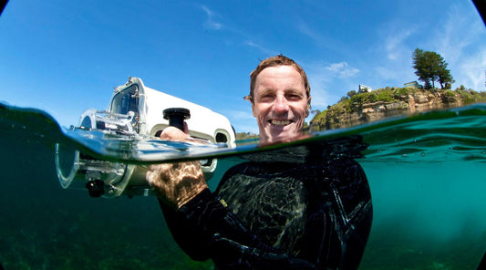 TIM BONYTHON HOLDING A CAMER IN A WATERPROOF CASING IN THE WATER SMILING AT THE CAMERA.