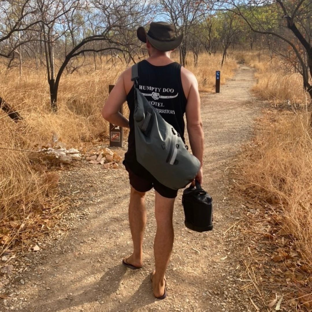 A MAN WALKING DOWN A DRY BUSH TRACK WEARING HIS NORTH STORM WATERPROOF DRY BAG OVER HIS SHOULDER IN AUSTRALIA..