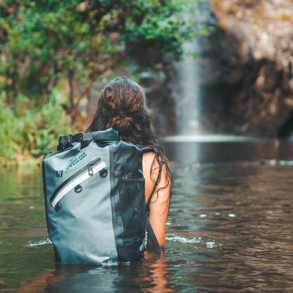 A lady wading waist deep in water wearing a 30L Waterproof Bag from North Storm®  and heading toward a beautiful waterfall.